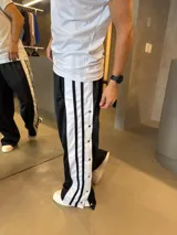 Pantalón deportivo negro de corte oversize con franjas laterales blancas y botones a presión a lo largo de las piernas.