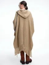 Ruana tejida de color beige con capucha integrada y terminaciones con flecos en el ruedo. Presenta un diseño envolvente y holgado, confeccionada en una mezcla de lana merino extrafina y viscosa.