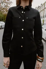 Camisa entallada color marrón oscuro, con cuello camisero y manga larga. Presenta dos bolsillos de plastrón en el delantero y cierre frontal con botones a presión dorados.