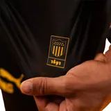 Camiseta de fútbol Puma del Club Atlético Peñarol, edición aniversario manga larga. Diseño con mitad derecha a rayas verticales amarillas y negras, y mitad izquierda negra con escudo retro "CUR" en dorado. Cuenta con cuello tipo polo negro con detalles amarillos y sponsors impresos en el frente y mangas.