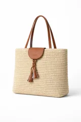 Bolso tipo tote bag de rafia tejida color beige, con asas y solapa superior de cuero marrón. Incluye un colgante decorativo con borlas de cuero.