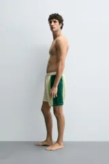 Bañador de hombre color blanco con diseño de bandas verticales en tonos de verde, simulando un efecto tie-dye o acuarela. Posee cintura ajustable con cordón, bolsillos laterales y forro interior.