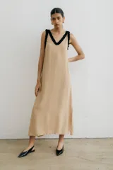 Vestido largo color beige claro, de corte recto y holgado, con escote en V profundo ribeteado en negro con volados. Presenta tirantes finos con el mismo detalle de volado negro.