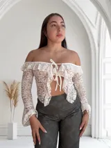 Top de encaje blanco con mangas largas acampanadas, hombros descubiertos, volados y lazo ajustable en el frente.