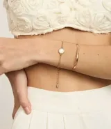 Set de dos pulseras doradas: una de cadena fina con dije circular de resina blanca y otra rígida.