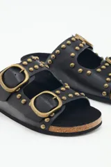 Sandalia plana negra de estilo 'dos hebillas' con tachas metálicas doradas decorando las tiras y hebillas ajustables doradas. La plantilla es de corcho y la suela de EVA.