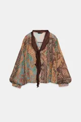 Blusa de manga larga con escote en pico y detalle de lazo frontal. Presenta un diseño estampado estilo paisley en tonos tierra, verdes y rojizos, con corte holgado y puños ajustados.
