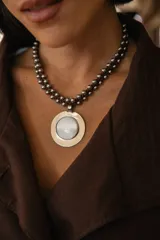 Collar corto con dije de medallón circular de nácar engarzado en metal plateado. La cadena está compuesta por un tiento de gamuza trenzado con cuentas metálicas esféricas.