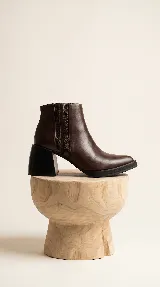 Bota corta de cuero marrón con diseño de paneles laterales texturizados. Presenta un taco ancho cuadrado en color negro y suela de plataforma baja, ofreciendo un estilo moderno y estable.
