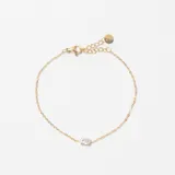 Pulsera de cadena fina dorada con un pequeño cristal central transparente. Cuenta con cierre de mosquetón y cadena de alargue con una pequeña placa circular.
