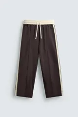 Pantalón tipo jogger de corte wide fit, confeccionado en tejido compacto color marrón oscuro. Presenta cintura elástica con cordón ajustable, bolsillos frontales y bandas laterales en contraste color beige. Incluye detalle de bolsillo trasero de vivo.