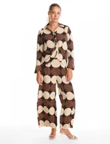 Conjunto de dos piezas compuesto por camisa de manga larga con nudo frontal y pantalón estilo palazzo de pierna ancha. Ambas prendas presentan un estampado geométrico de círculos grandes en tonos marrón, negro y beige. La camisa cuenta con cuello solapa y cierre frontal con botones, mientras que el pantalón posee tiro alto y corte holgado.