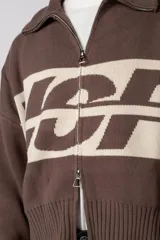 Campera tejida de punto con cierre frontal completo y cuello polo. Presenta un diseño de franja horizontal ancha en color crema con estampado gráfico oscuro sobre una base de color marrón. Posee puños y ruedo acanalados, con un pequeño parche de marca en el puño izquierdo.