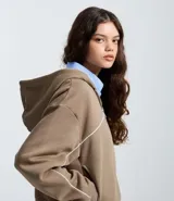 Saco con capucha de corte holgado, confeccionado en mezcla de algodón y poliéster color marrón. Presenta cierre frontal por cremallera, puños y ruedo acanalados, y detalle de piping blanco en contraste a lo largo del pecho y las mangas.