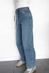 Pantalón de jean estilo mom fit en tono azul clásico, con diseño de tiro alto y lazo ajustable en la cintura. Presenta un corte relajado y holgado en las piernas, confeccionado en denim rígido con estructura.