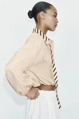 Camisa de popelín color beige con cuello solapa y lazada frontal en contraste con estampado de rayas marrones. Presenta mangas largas abullonadas con puños elásticos, bolsillos con solapa en el pecho y bajo elástico.