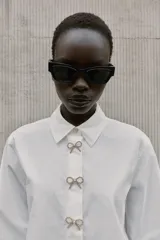 Camisa de popelín blanca con cuello solapa y manga larga. Presenta detalle de botones joya decorativos en forma de lazo en la parte frontal y cierre oculto por solapa.