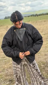 Campera puffer negra con diseño matelaseado en rombos, silueta amplia y corte bomber. Presenta cuello alto, cierre frontal y un detalle triangular de cuero en la parte superior con logo grabado. Incluye mangas largas con puños elásticos y bolsillos laterales funcionales.