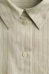 Camisa de corte oversized, color beige claro con rayas finas, cuello clásico, cierre frontal con botones, un bolsillo aplicado en el pecho, hombros caídos y mangas largas con puños abotonados.