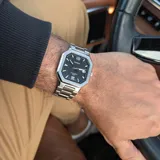 Reloj analógico Casio con caja octogonal de acero inoxidable y malla de eslabones plateada. Presenta una esfera negra con textura estriada, índices aplicados, números 6 y 12, manecillas luminiscentes y ventana de calendario a las tres.