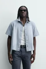 Camisa de corte boxy fit confeccionada en denim ligero de algodón. Presenta cuello solapa, manga corta y cierre frontal con botones. Diseño con efecto lavado.