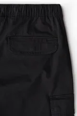 Pantalón cargo negro de corte holgado, con cintura elástica ajustable con cordón, bolsillos laterales al bies, bolsillos tipo cargo con solapa y botón a presión en las piernas y bolsillos traseros con solapa y botón a presión. Cuenta con pliegues cosidos en las rodillas y cordón ajustable oculto en el bajo.