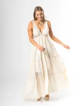 Vestido largo color crudo, confeccionado en mezcla de tencel y lino, con escote en V pronunciado, breteles anchos terminados en lazos en los hombros, y falda larga de varios niveles con volados amplios y fluidos.
