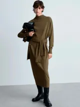 Vestido midi de punto fino en color verde oliva, con cuello alto tipo perkins, manga larga y diseño recto. Presenta un detalle de lazo anudado en la cintura y terminaciones en canalé.