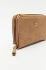 Billetera de formato rectangular con cierre perimetral, confeccionada en material símil cuero de textura suave. Presenta un aplique metálico circular con logo grabado en el frente y tirador de cierre en el mismo material.