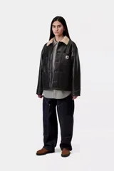 Chaqueta de trabajo Carhartt WIP modelo Michigan Shepton, color negro, confeccionada en piel sintética con cuello y forro de corderito sintético en tono tostado. Presenta cierre frontal de botones, cuatro bolsillos delanteros de parche (uno con solapa) y etiqueta cuadrada con el logo de la marca.