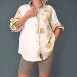Camisa blanca con estampado de hojas en tonos beige y verde, cuello camisero, mangas largas con lazos en los puños y botones de madera.