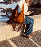 Bolso marrón de gamuza con flecos largos, asa de hombro y detalle de pasamanería con nudos.