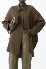 Camisa larga de manga larga, color marrón oscuro, con cuello camisero y botones frontales. Presenta un corte holgado y frunce sutil en las mangas.