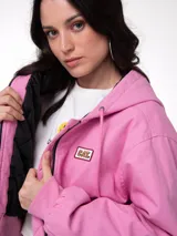 Chaqueta de lona color rosado con capucha ajustable, cierre frontal con cremallera y forro interior acolchado con diseño de diamantes. Presenta puños y bajo acanalados, bolsillos laterales y parche con el logo de la marca en el pecho.