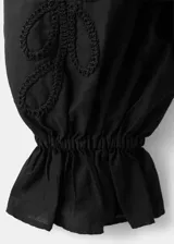 Blusa de algodón negra con detalles florales bordados, cuello en pico con cierre de lazo, manga larga abullonada y diseño recto con vuelo en la cintura.