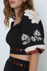 Top corto negro con escote bardot, mangas cortas abullonadas con bordado floral blanco y detalle de recortes con tiras horizontales color café en la cintura.