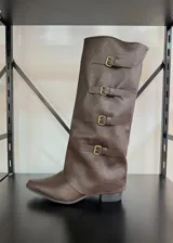 Bota de caña alta estilo slouchy color marrón, confeccionada en material sintético con acabado mate. Presenta un diseño con cuatro correas horizontales decorativas con hebillas metálicas doradas y un taco bajo cuadrado.
