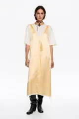 Vestido midi de diseño bimateria con sobrevestido de satén color amarillo claro. Presenta escote en V profundo con lazo frontal, detalles de encaje en el ruedo y aberturas laterales con cordones. Se combina con una base de remera blanca de manga corta y cuello redondo.