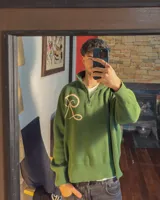 Sweater tejido de algodón en color verde oscuro, con cuello alto y cierre frontal parcial. Presenta un diseño de letra R bordada en color beige en el frente.