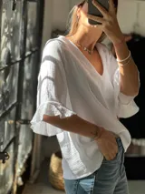 Blusa de muselina blanca con escote en V y mangas cortas con volados en los puños. Presenta una textura arrugada característica del tejido.