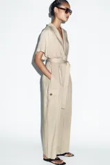 Mono largo de cuello solapa y escote en pico cruzado, con mangas cortas. Presenta bolsillos laterales de plastrón con solapa y botones, y un cinturón del mismo tejido para ajustar en la cintura. Cierre frontal con cremallera y botón interior.