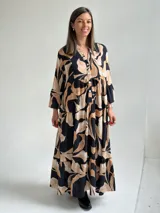 Vestido largo negro con estampado de hojas en tonos beige y tostado, de corte amplio y fluido, con escote en V y botonadura frontal parcial. Cuenta con mangas largas y volados en el faldón.
