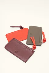 Monedero rectangular de cuero vacuno con textura granulada en color marrón topo. Cuenta con cierre superior metálico y un tirador decorativo de cuero en color naranja con un nudo. Presenta un pequeño logo grabado en el centro.