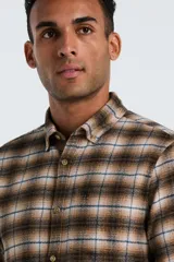 Camisa de manga larga confeccionada en tela de franela con estampado de cuadros en tonos marrones, beige y líneas azules. Presenta cuello clásico abotonado, cierre frontal con botones y un bolsillo pequeño en el pecho.