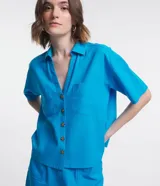 Camisa de manga corta confeccionada en mezcla de viscosa y lino color turquesa. Presenta cuello camisero con escote en V, bolsillos frontales de parche y cierre frontal con botones geométricos de efecto carey.