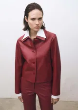 Chaqueta corta de cuero color rojo con cuello camisero blanco, cierre frontal con botones y mangas largas.