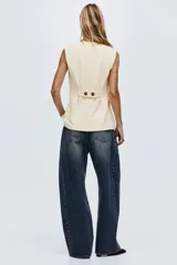 Chaleco largo color beige, con escote en V, cierre de botones a presión ocultos y bolsillos decorativos frontales. Presenta cintura entallada con pinzas y trabilla con botón en la espalda.