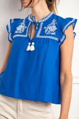 Blusa blanca de viscosa con bordados negros en contraste. Presenta escote en V con lazo y borlas, mangas cortas tipo volado y detalles bordados de flores y motivos geométricos en el pecho y hombros.