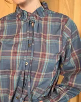 Camisa de manga larga con estampado de cuadros en tonos azules y rojizos. Presenta un cuello alto con volados y puños elásticos con detalle de volado en el extremo. Cierre frontal con botones ocultos.