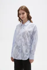 Camisa de algodón con diseño de rayas verticales finas en color celeste y blanco, con apliques florales bordados en relieve. Presenta cuello camisero, cierre frontal con botones y mangas largas con puños abotonados.